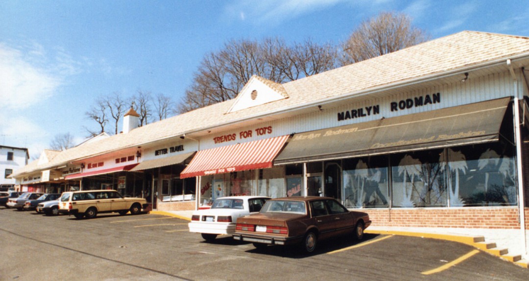 Historical view of row of retail stores with 1980s cars in parking spaces